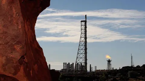 Getty Images A red rock shelf in the foreground with an industrial site in the background, including a flaming gas tower
