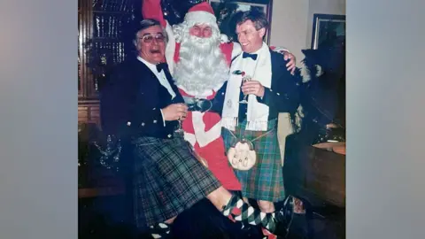 Garry Crockett Two men wearing kilts pose for a photo with another man dressed as Santa