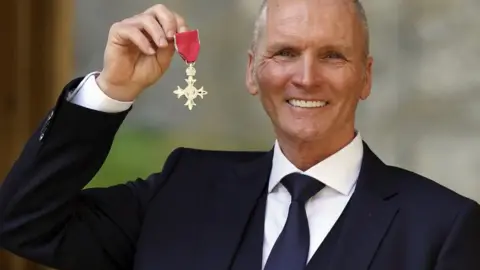 PA Media Thomas is holding is MBE in 2021. He is wearing a dark suit and tie and smiling at the camera.