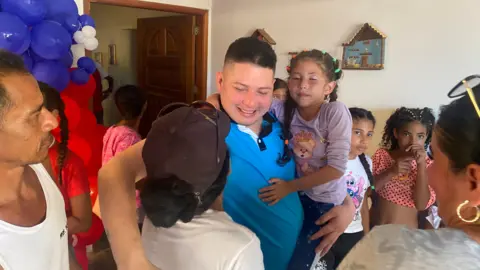 Gustavo Ocando Andy Perozo, wearing a blue polo shirt and smiling, carries a young girl on his arm and embraces a woman. Other children look. In the background, balloons can be seen