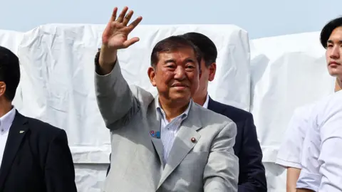 Reuters Japan's Prime Minister Shigeru Ishiba, who is also the leader of the ruling Liberal Democratic Party (LDP), waves to voters from atop an election campaign van during the LDP's election campaign tour for the July 20, 2025 Upper House election, in Yokohama, south of Tokyo, Japan July 18, 2025. He is wearing a light grey suit with no tie, and has a slight smile on his face. 