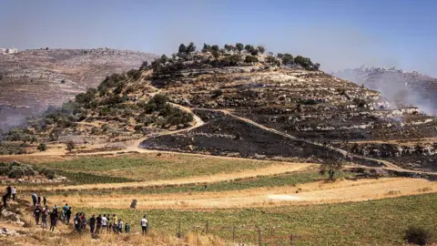 AFP File photo showing Palestinians looking on as a fire burns on a hilltop that was seized by Israeli settlers near the town of Sinjil, in the occupied West Bank (4July 2025)