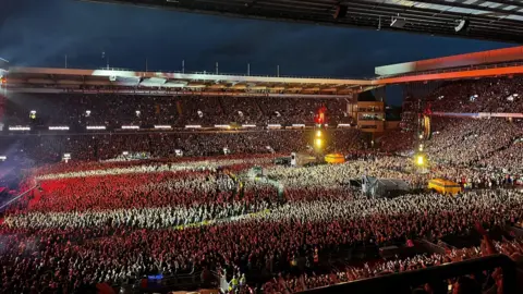 A huge sea of people are seen in the stadium, lit up by red lights from the stage