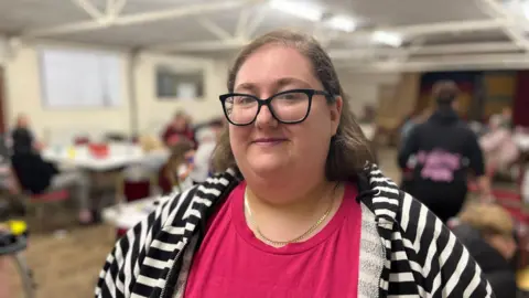 Sophie smiles at the camera from inside the community centre. She wears black-framed glasses, a bright pink top and a black and white striped hoodie. 