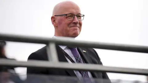 Getty Images John Swinney, who wears a dark suit with a tartan tie, arrives on a golf course. He is bald, wears glasses and is smiling in the picture. Swinney is sitting down.