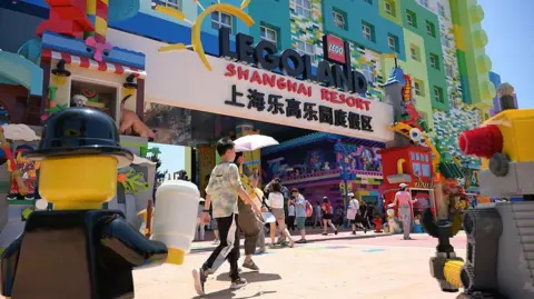 Getty Images Dozens of visitors stream into Legoland Shanghai through an entrance at the base of a colourful hotel. In the photo is a large banner displaying the words "Legoland Shanghai Resort". Large mascots, based on Lego minifigurines, are pictured at the foreground of the shot. 