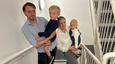 A mum and dad carrying one child each standing in a building stairwell. 