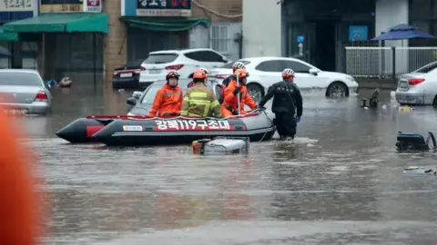 Reuters Firefighters on a rescue boat navigating through a flooded neighbourhood caused by torrential rain in Daegu, South Korea. Cars are parked along the street and some furniture, including swivel chairs, can be seen floating in the floodwaters.