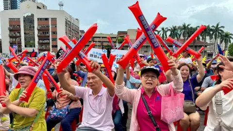 BBC / Tessa Wong Crowds of people seated at a daytime anti-recall rally in New Taipei hold up large red inflatable sticks, crossing them to indicate their opposition to the Great Recall. In the foreground you can see a woman wearing a pink polo shirt and pink cardigan carrying a pink bag, and a man wearing a light polo shirt. In the background you can see municipal buildings and trees.