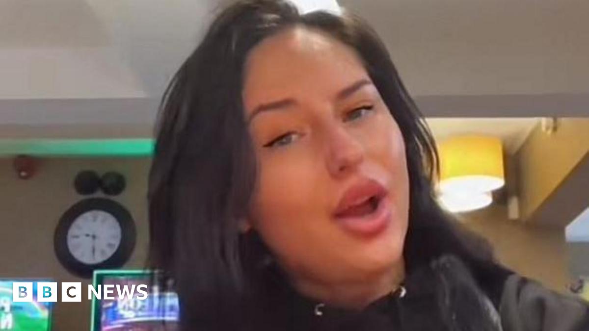 A woman with long dark hair poses in a pub, with her mouth open and a clock in the background.