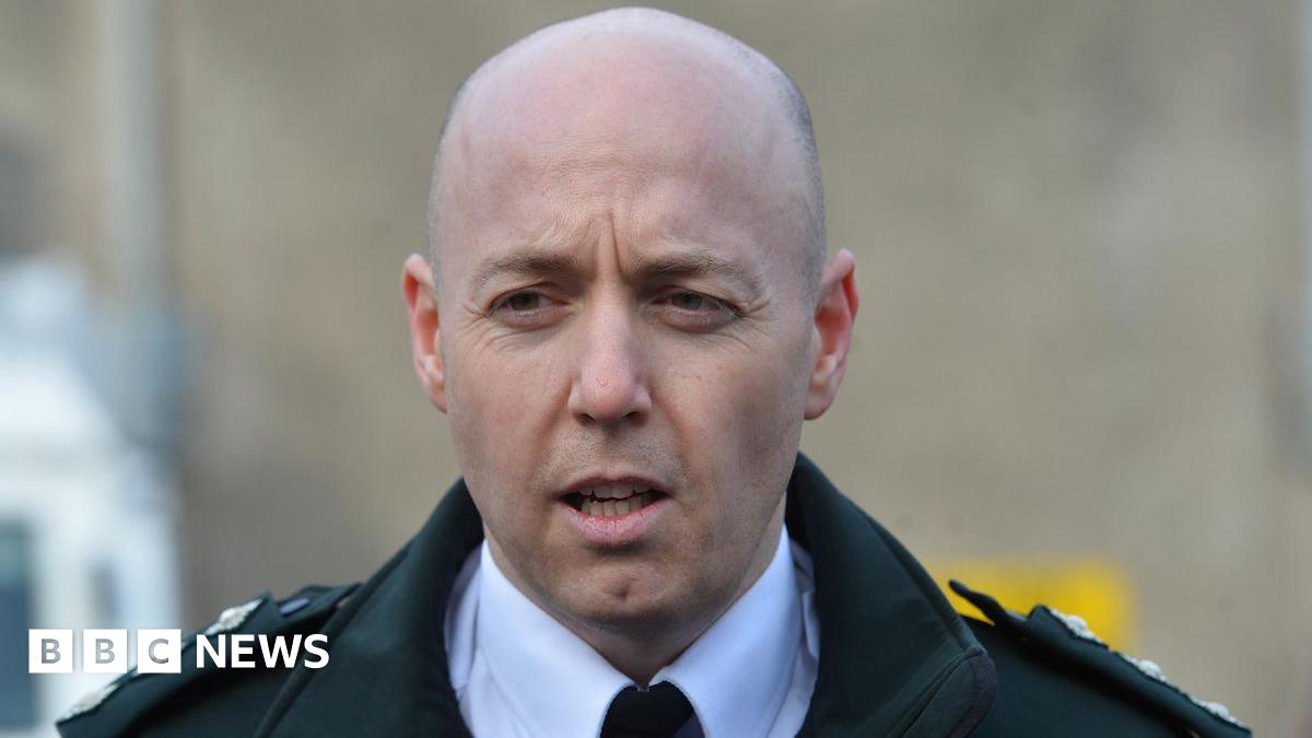 A man sits in front of a mic. He's wearing a suit. Wooden panelling is on the walls behind him.