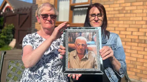 BBC Two women hold up a silver framed picture of an older man.
The woman on the left, Susan Holmes, is wearing a black and white floral patterned top and dark glasses and has short blonde hair while the woman on the left, Lisa Jones, has long dark hair and is wearing a denim jacket and glasses. The man has short white hair and is looking directly at the camera while wearing a striped shirt. Behind them you can see the wall and window of a house.