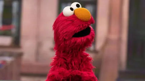 Getty Images Elmo on the Sesame Street set. Elmo is a furry red Muppet with a large orange nose and big round eyes. He looks up to the right of the frame, with the set blurred behind him. 