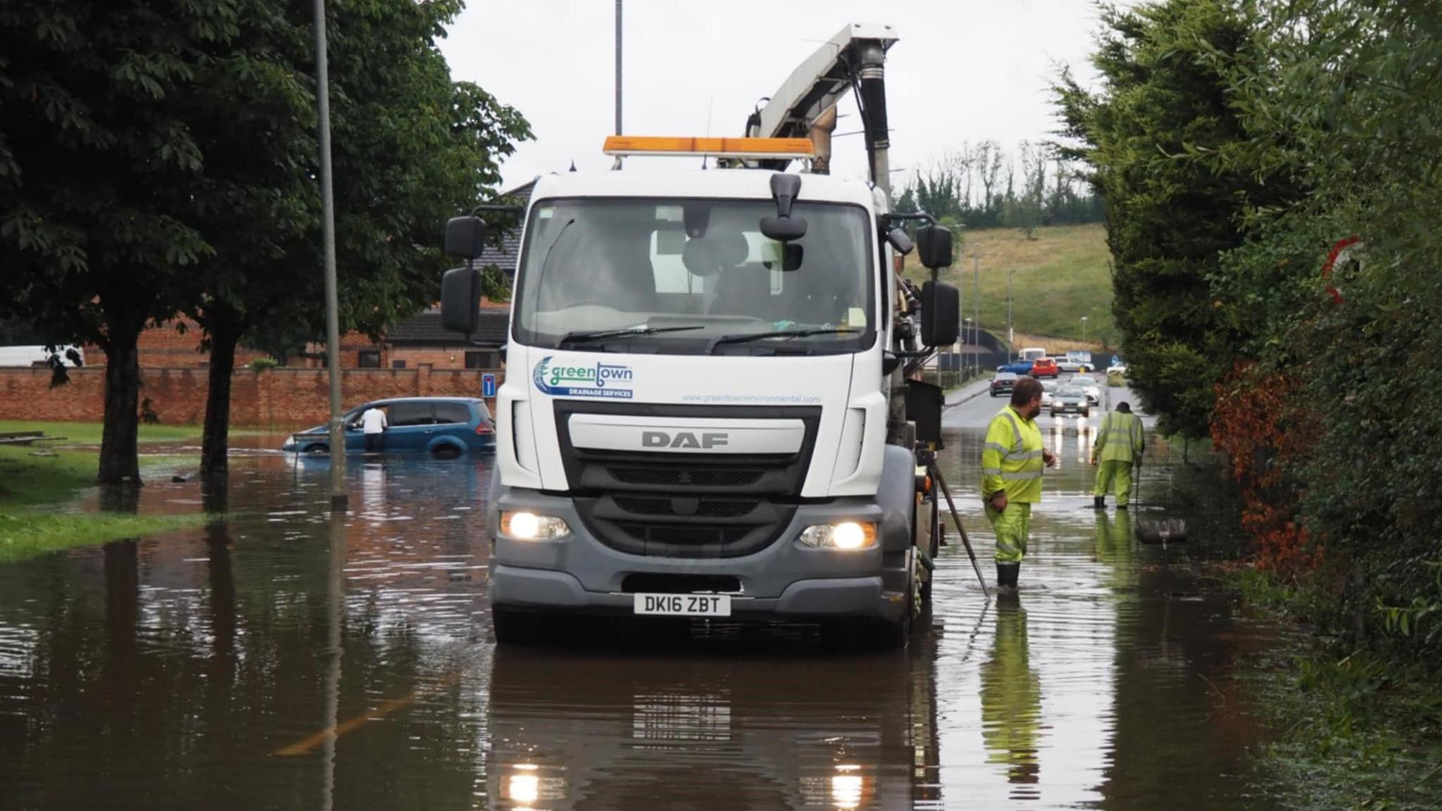 A wide view of the flooded Dublin Road in Enniskillen. A large white truck with a Greentown Drainage Services sign is parked in the middle of the road. Two men in yellow high-viz suits and wellies are standing ankle-deep in the water.  IN the distance, a man leans into a blue hatchback car which is submerged up to its bumper in the floodwater.