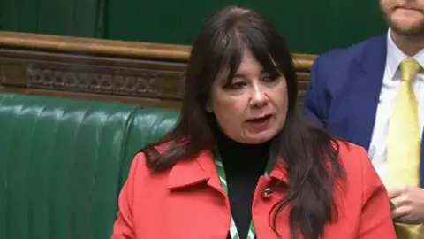 Parliament TV Anneliese Midgley, who has dark hair and is wearing a red jacket, is speaking in the House of Commons.