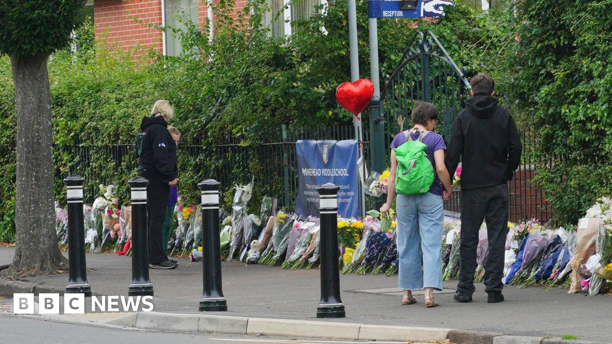 Four people stand with their backs turned outside Minehead Middle School. They are looking at bunch of flowers which have been lined up against the metal fence outside the school. There is a red balloon in the shape of a heart attached to the fence.