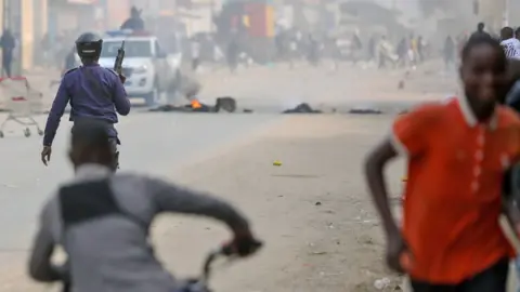 EPA A police officer moving towards protesters on a street in Luanda. Some of them can be seen in the hazy distance; in the foreground a protester in a red shirt runs towards the camera. A man on a bike can be seen swerving behind the officer. Debris can be seen burning on the road where a police vehicle can also be seen - 28 July 2025.