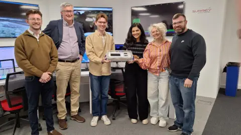 University of Surrey A group of six people stand in the Surrey Space Centre, with these words written on the wall behind them. Those in the middle are holding a pale plastic box which has a dark black rectangle in the middle of it.