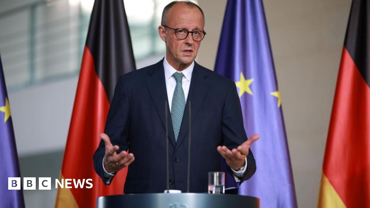 Merz gestures as he speaks at a lectern. A line of furled EU and German flags hang behind him