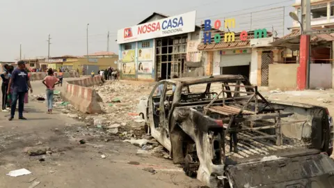 AFP/Getty Images A group of people look on at a looted supermarket with a broken shop front and rubbish strewn outside. To the right is a burnt out pick-up truck in Luanda's Kalemba 2 district - 29 July 2025.