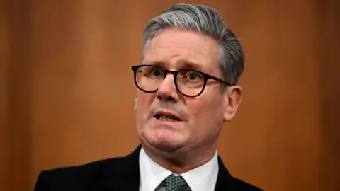 Reuters Sir Keir Starmer, dressed in a black suit jacket, white shirt, and green patterned tie, stands against a plain warm brown background. 