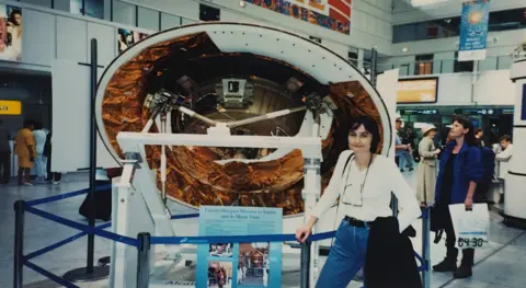 Michele Dougherty Michele Dougherty age 30 in a white top and blue jeans with glasses hanging around her neck and a black jacket slung over her arm. She stands in front of a round part of a spacecraft which is partly white but with copper coloured foil in parts. It is cordoned off by a blue ribbon rail. The scene appears to be in a museum, or perhaps and airport and other people are milling around.