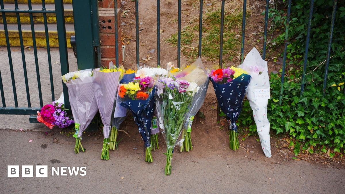 A row of about a dozen bouquets of flowers are resting against school gates.