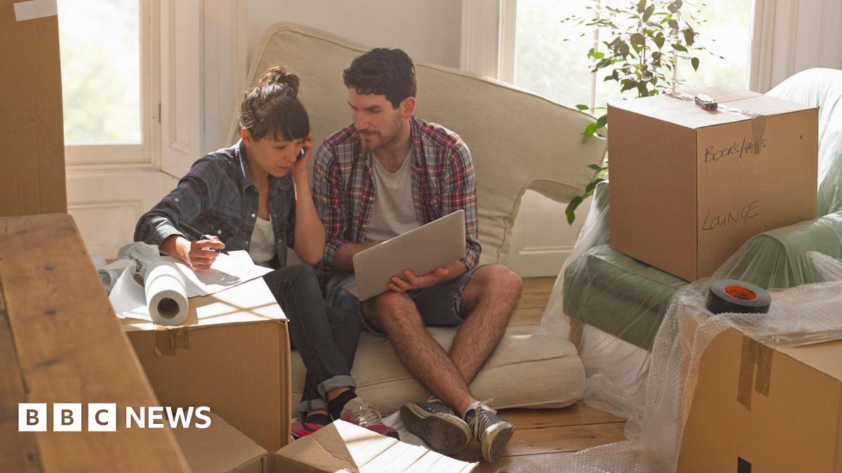 A man and a woman sit on a cushion looking at a laptop computer surrounded by unpacked boxes.