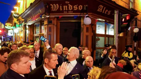 Reuters President Joe Biden outside a corner pub called The Windsor in a busy street in Ireland. he is surrounded by a large crowd and is taking a selfie with a phone in the street. He is smiling and dressed in a dark suit and tie with a plain white shirt.