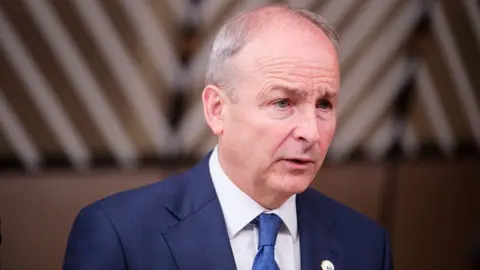 Getty Images A man looks and leans to his left. he is wearing a navy suit, white shirt and dark blue tie. He is balding and has grey at the side of his head. The background is blurred.