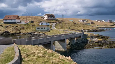 Getty Images The island communities of Housay and Bruray in the island group of Out Skerries. Houses are scattered across a grassed area with a bridge at the centre of the photograph connecting the two islands and the sea on the right.