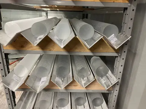 PNRA/IPEV 15 ice cores that look frosty and are about 15cm in diameter are inside metal gulleys. They are stacked on wood and metal shelves.