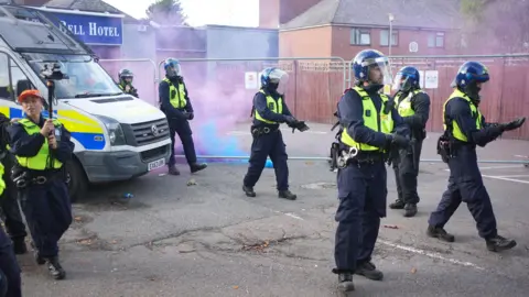 PA Media Several police officers in dark trousers, yellow high-visibility jackets and riot helmets stand outside The Bell Hotel in Epping. There is a police van on the left hand side of the photo, and a female police officer holding a camera on a monopod. There is blue and purple smoke in the background, and the hotel is blocked off by metal fencing.