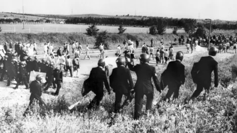 PA Media A black and white image of miners and police officers with riot gear walking along a field. 