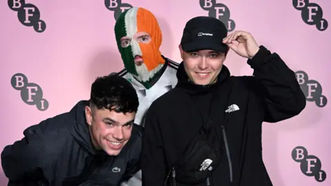 Getty Images Three young men stand together. One is wearing a green, white and orange balaclava. A pink backdrop is behind them.