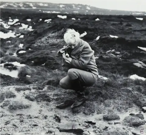 Longtail Films A black and white photograph shows Myra Hindley crouching on a rocky, grassy terrain. She is wearing a coat and boots, and is holding a small dog close to her chest. Snow patches are visible in the background.