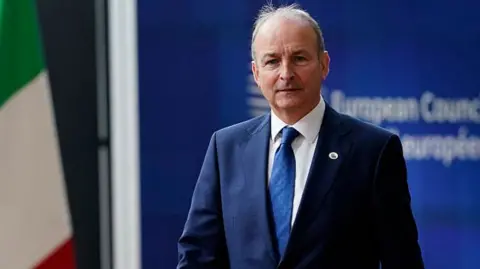 Getty Images A man looks into the camera. He has grey hair at the sides and is balding on top. He is wearing a navy suit, blue tie and white shirt. He is standing in front of blue signage with blurred white writing on it. There is an Italian flag to the man's right.