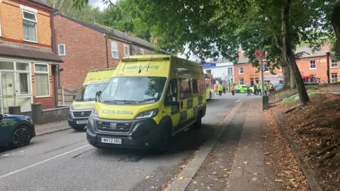 Richard Stead/BBC Two yellow ambulances are parked in the middle of a street. People can be seen walking along the street in the background. 