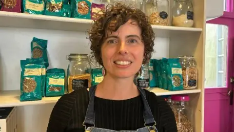 Gemma Beere smiles at the camera. She is wearing a denim apron and is stood in front of shelves filled with gluten free pastas and grains