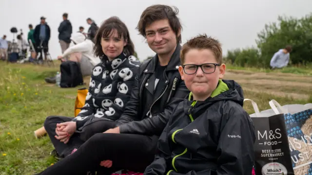 Abbie, Kevin and Cooper are dressed in black coats and sit on a red blanket with an M&S paper bag