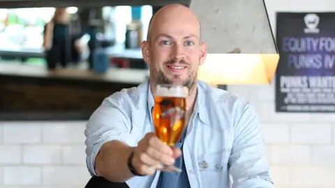 Getty Images A smiling bald, bearded man wearing a light blue shirt - with the sleeves rolled up - and a light blue t-shirt raises a full beer glass with his right hand. He has been photographed in a pub setting, with a white wall and a bar visible in the blurry background.