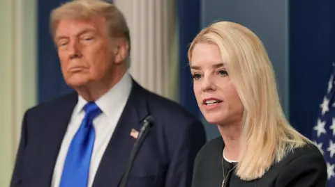 Reuters US Attorney General Pam Bondi, a woman with short blonde hair wearing a black blouse, speaks in front of a US flag, with US President Donald Trump behind  her wearing a dark blue jacket, white shirt and bright blue tie.