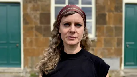Ellena Wood with long curly hair, a head scarf and a black top stands  in front of a small orange-brick chapel building.