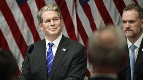 EPA US Treasury Secretary Scott Bessent smiles sitting in front of a microphone and a backdrop of American flags while US Trade Representative Jamieson Greer is in the background