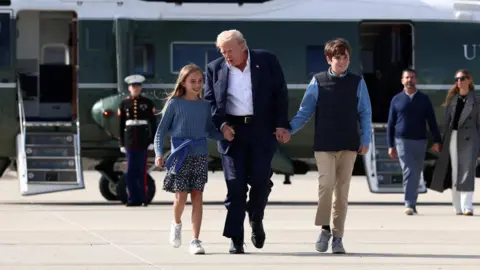 PA Media Trump walks from his helicopter Marine One to the presidential plane Air Force One with his grandchildren. He is holding their hands and talking to his granddaughter. He is wearing a dark suit and a white shirt