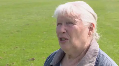 Iris Ritchie looking slightly off camera. She has white hair and is wearing a blue jacket with a grey collar.