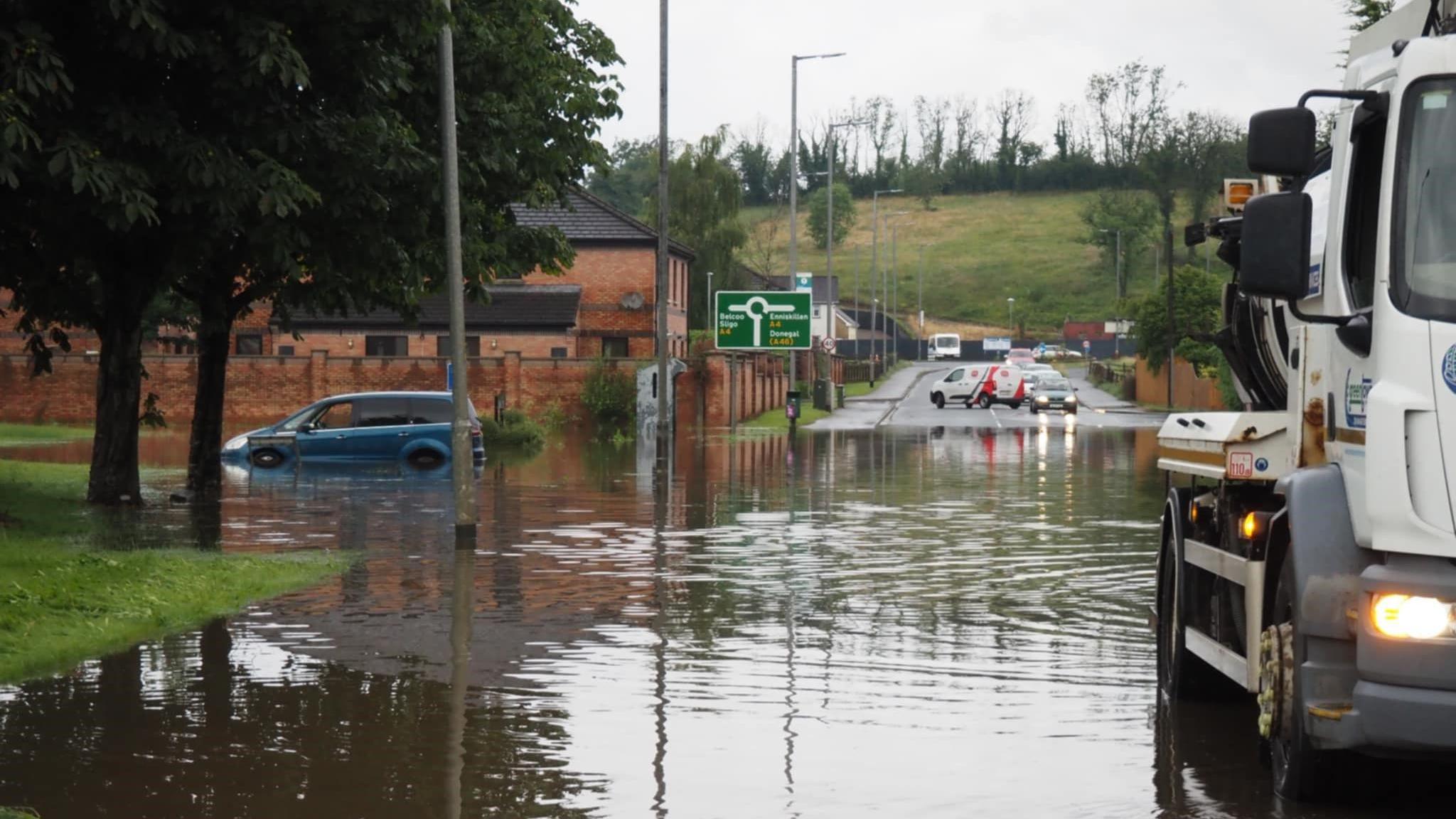 Flooding on the main Enniskillen to Dublin Road in Enniskillen on Sunday evening.  Part of the road is submerged in flood water. One blue hatchback car is parked on the left of the photo.  The water level is up to its rear bumper and more than half way up its rear wheel arch. A large white truck used to clear drains is in the foreground and cars are turning away from the area in the background.