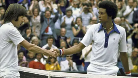 Sports Illustrated/Getty Images Jimmy Connors and Arther Ashe - both in tennis whites - shake hands over the net after Ashe's victory in the Wimbledon final of 5 July 2025. The clapping crowd can be seen behind them