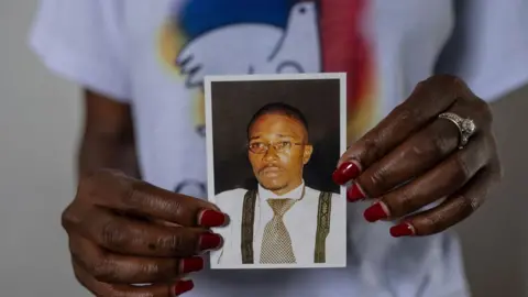 AP A close-up of a woman's hands, with read nail varnish, holding up a small photo of Floribert Bwana Chui Bin Kositi. In it he is staring ahead and looking serious. He is wearing oval wire-rimmed glassed, a silver and black dog tooth design tie, black braces and a white shirt. The woman's T-shirt, showing a peace dove design, can be seen in the background.