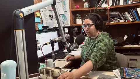 BBC Desire Hagan, wearing a green sweatshirt, glasses and earphones, speaks into a microphone in a radio studio.
